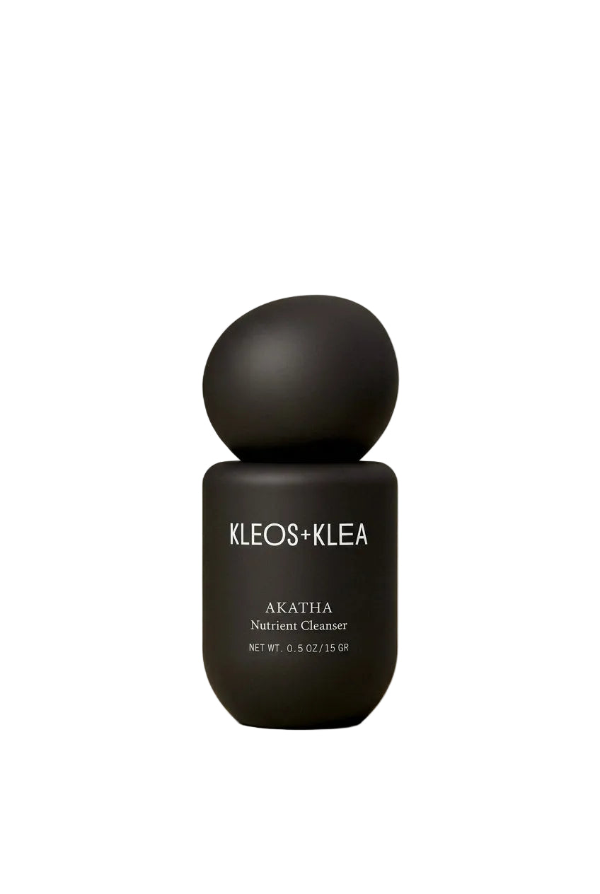 A matte black skincare bottle with a spherical cap, labeled 'KLEOS+KLEA AKATHIA Nutrient Cleanser', centered against a warm beige gradient background.