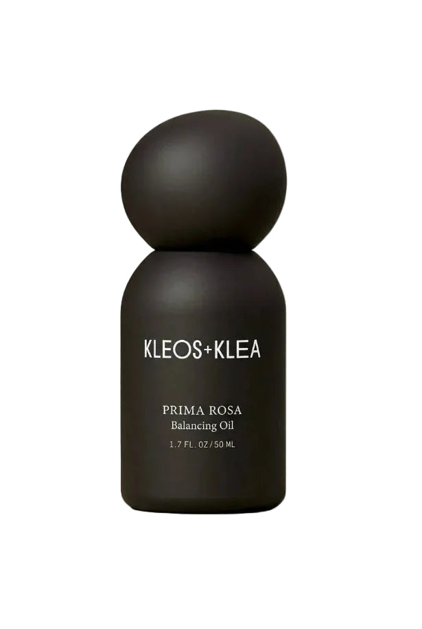 A matte black bottle of KLEOS+KLEA Prima Rosa Balancing Oil, featuring a distinctive rounded, double-bulb cap, set against a warm beige studio background.