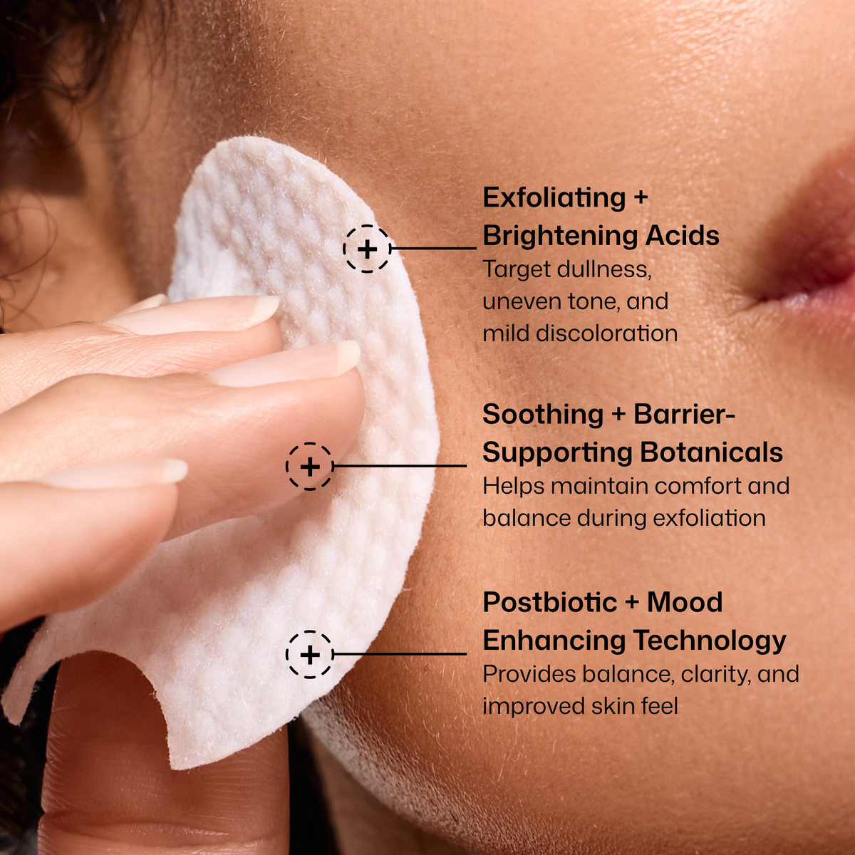 Close-up of a person's face with a white round cleansing pad being held against the cheek, with three text callouts describing skincare benefits: exfoliating and brightening acids, soothing and barrier-supporting botanicals, and postbiotic + mood enhancing technology.