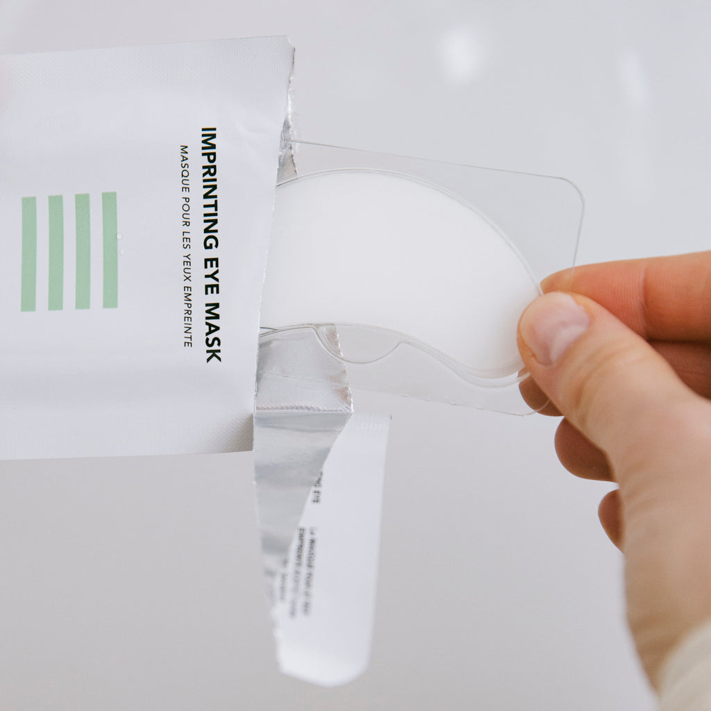 Hand holding an eye mask with packaging labeled 'Imprinting Eye Mask' on a white background